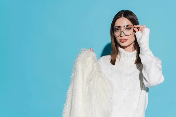 Stylish young woman adjusting trendy eyeglasses and holding white faux fur jacket on blue — Stock Photo