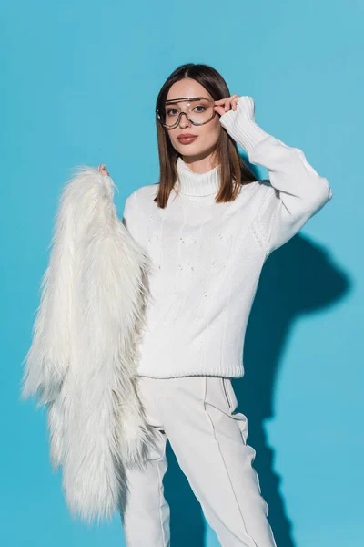 Pretty and young woman adjusting trendy eyeglasses while holding white faux fur jacket on blue — Stock Photo