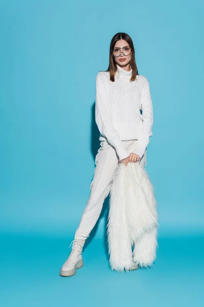 Full length of stylish young woman in trendy eyeglasses and turtleneck holding white faux fur jacket on blue — Stock Photo