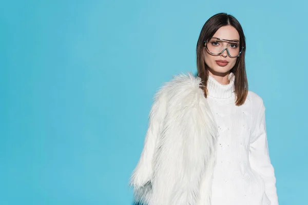Stylish young woman in trendy eyeglasses and turtleneck holding white faux fur jacket on blue — Stock Photo