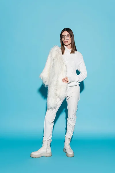 Comprimento total da mulher elegante em óculos da moda e gola alta branca segurando jaqueta de pele de falso branco no azul — Fotografia de Stock