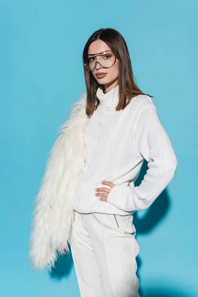 Elegante donna in occhiali da vista e dolcevita bianco in piedi con mano sul fianco mentre tiene giacca di pelliccia sintetica su blu — Foto stock