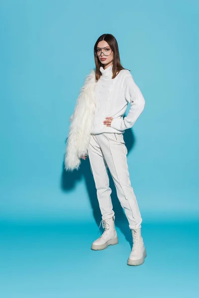 Full length of stylish woman in eyeglasses and turtleneck standing with hand on hip while holding faux fur jacket on blue — Stock Photo