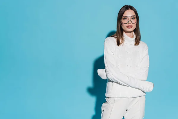 Pretty young woman in trendy eyeglasses and white turtleneck posing with crossed arms on blue background — Stock Photo