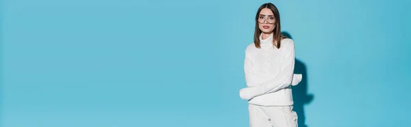 Bella giovane donna in occhiali alla moda e dolcevita bianco in posa con braccia incrociate su sfondo blu, banner — Foto stock