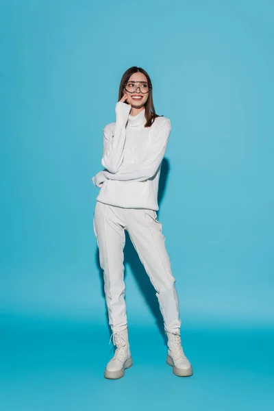 Longitud completa de alegre mujer joven en gafas y traje totalmente blanco posando en azul - foto de stock