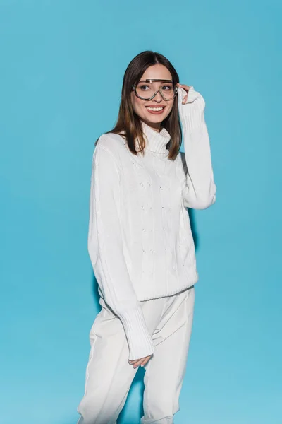 Fröhliche junge Frau in trendiger Brille und komplett weißem Outfit posiert auf blauem Grund — Stockfoto