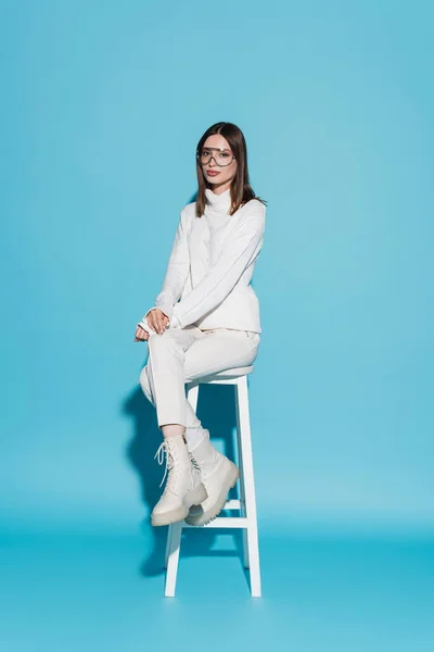 Полная длина молодой и стильной женщины в полном белом наряде, сидящей на стульчике на синем — стоковое фото