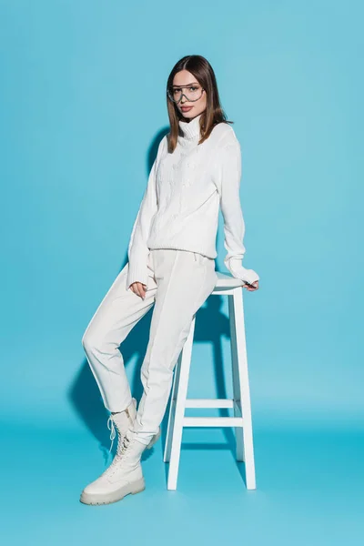 Volle Länge der jungen und stilvollen Frau in weißem Outfit lehnt auf Hochstuhl auf blau — Stockfoto
