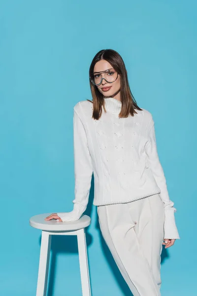 Trendy model in white outfit and eyeglasses leaning on high chair on blue — Stock Photo
