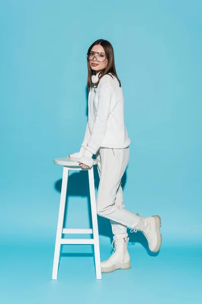 Повна довжина модної моделі в білому вбранні та окулярах, що спираються на високий стілець на синьому — стокове фото