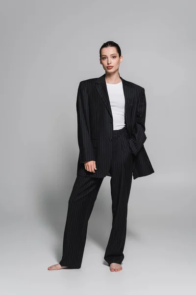 Pleine longueur de jeune femme élégante en costume noir posant avec la main dans la poche sur le gris — Photo de stock