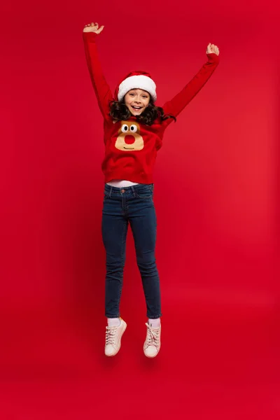 Full length of happy girl in santa hat and sweater levitating on red — Stock Photo
