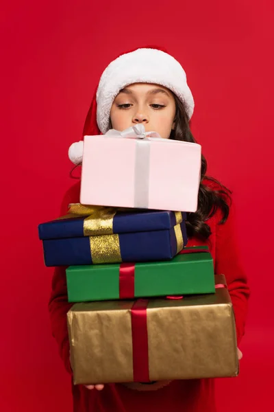 Девушка в шляпе Санты и свитере с кучей рождественских подарков, изолированных на красный — стоковое фото