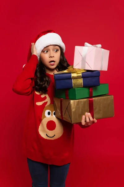 Шокированная девушка в шляпе Санты и свитере держит кучу рождественских подарков, изолированных на красный — стоковое фото