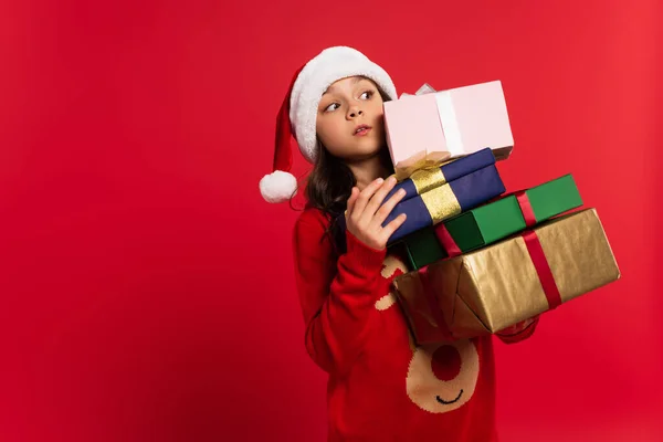 Surprised girl in santa hat and sweater holding bunch of Christmas presents isolated on red — Stock Photo