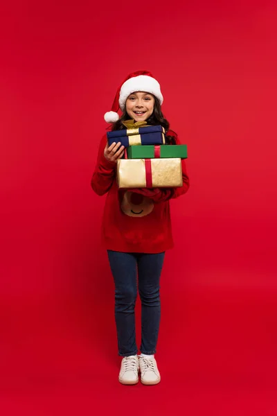 Full length of happy girl in santa hat and sweater holding bunch of Christmas presents on red — Stock Photo