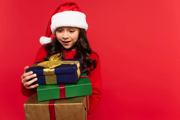 Happy girl in santa hat and sweater holding bunch of Christmas presents isolated on red — Stock Photo