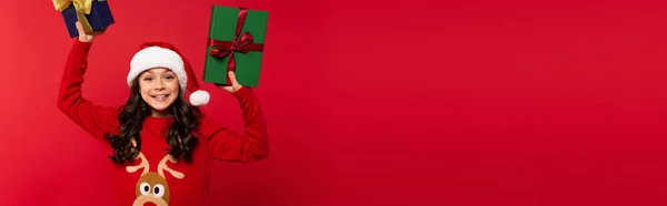Happy girl in santa hat and sweater holding Christmas presents isolated on red, banner — Stock Photo