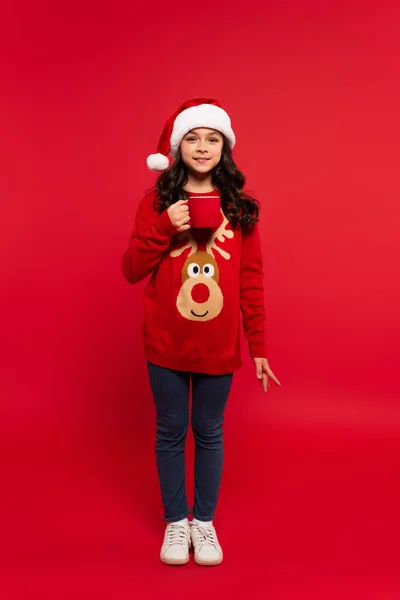 Longitud completa de la chica feliz en sombrero de santa y suéter taza de cacao en rojo - foto de stock