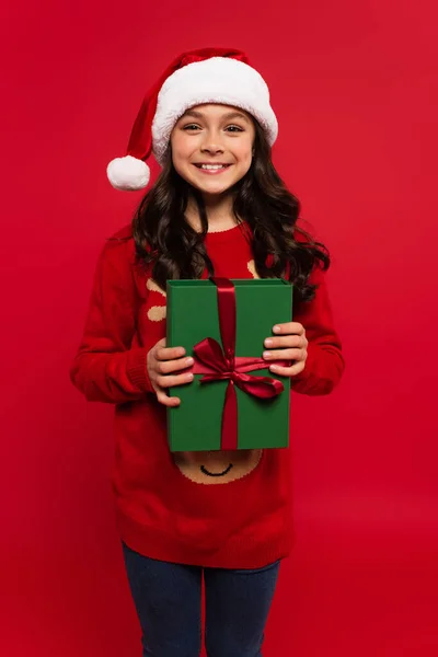 Happy girl in santa hat and knitted sweater holding wrapped Christmas gift box isolated on red — Stock Photo