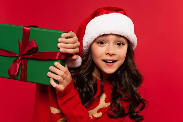 Amazed girl in santa hat and sweater holding wrapped Christmas gift box isolated on red — Stock Photo