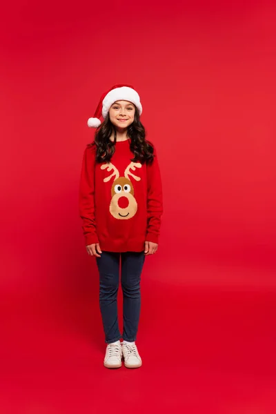 Pleine longueur de fille heureuse en pull de Noël et chapeau de Père Noël debout sur rouge — Photo de stock