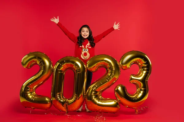 Longitud completa de la chica excitada en suéter de pie cerca de los globos con 2023 números en rojo - foto de stock