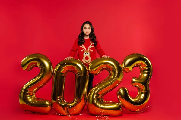 Повна довжина щасливої дівчини в светрі, що стоїть біля кульок з 2023 номерами на червоному — стокове фото