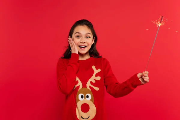 Happy girl in Christmas sweater holding shiny sparkler isolated on red — Stock Photo
