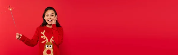Fille heureuse en pull de Noël tenant brillant brillant isolé sur rouge, bannière — Photo de stock
