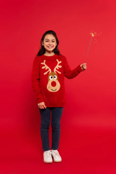 Full length of cheerful girl in Christmas sweater holding shiny sparkler on red — Stock Photo