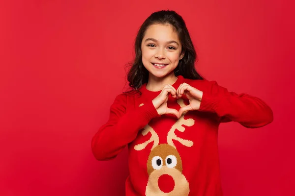 Niño alegre en suéter de invierno mostrando el signo del corazón con las manos aisladas en rojo - foto de stock