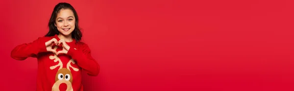 Enfant gai en pull d'hiver montrant signe du coeur avec les mains isolées sur rouge, bannière — Photo de stock