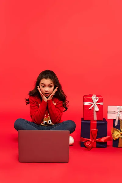 Schockiertes Kind in Winterpullover und Jeans sitzt neben Laptop und Weihnachtsgeschenken auf rotem Grund — Stockfoto