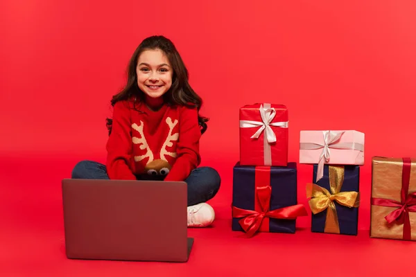 Pleine longueur de gamin joyeux en pull d'hiver et jeans assis près d'un ordinateur portable et cadeaux de Noël sur rouge — Photo de stock