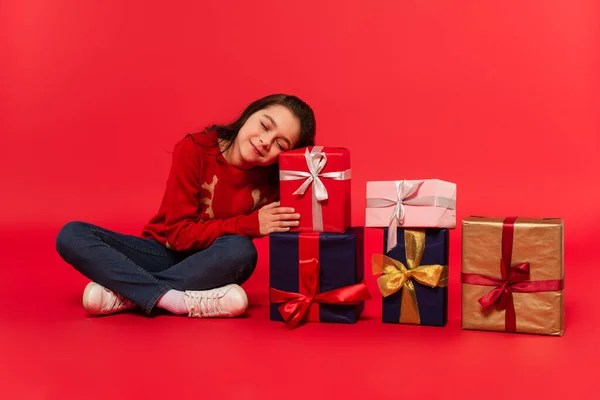 Full length of pleased kid in winter sweater and jeans sitting near Christmas presents on red — Stock Photo