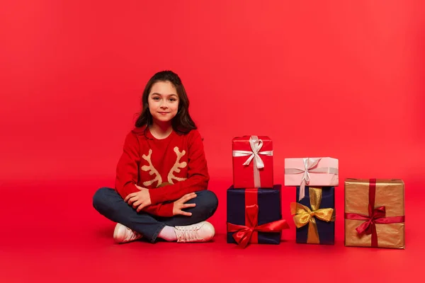 Повна довжина веселого дитини в зимовому светрі і джинсах, що сидять біля різдвяних подарунків на червоному — стокове фото