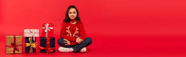Comprimento cheio de criança alegre em suéter de inverno e jeans sentados perto de presentes de Natal no vermelho, bandeira — Fotografia de Stock
