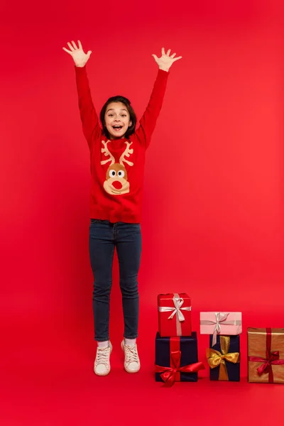 Tutta la lunghezza del bambino eccitato in maglia maglione gesticolando vicino regali di Natale sul rosso — Foto stock