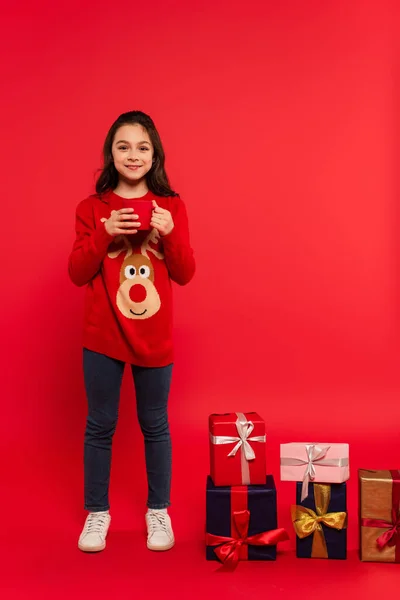 Longitud completa de niño feliz en suéter de punto sosteniendo taza de cacao cerca de regalos de Navidad en rojo - foto de stock