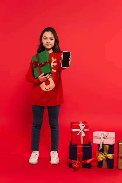 Longitud completa de niño feliz en suéter de punto que muestra el teléfono inteligente con pantalla en blanco cerca de los regalos de Navidad en rojo - foto de stock
