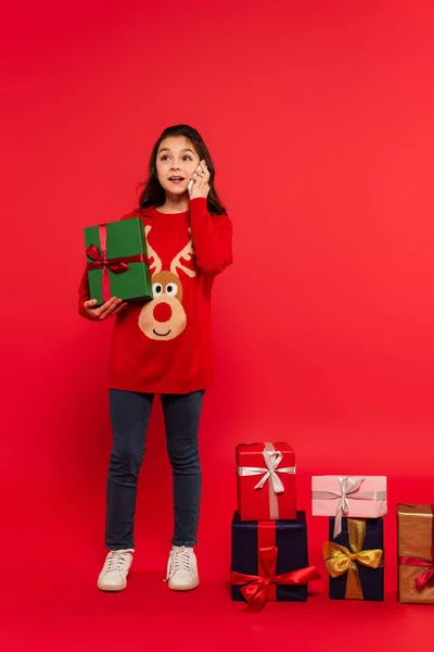 Tutta la lunghezza del bambino felice in maglia maglione parlando su smartphone vicino regali di Natale sul rosso — Stock Photo
