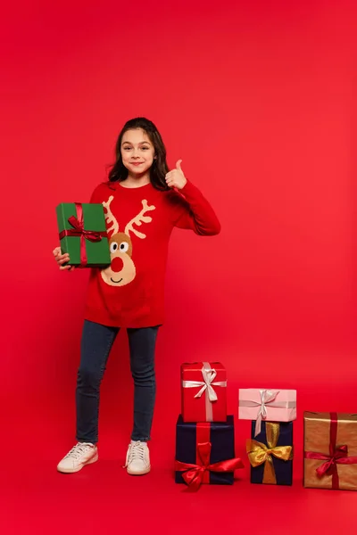 Повна довжина щасливої дитини в в'язаному светрі, що показує великий палець біля різдвяних подарунків на червоному — стокове фото