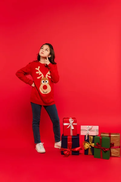Nachdenkliches Kind in Strickpullover posiert mit Hand auf Hüfte bei Weihnachtsgeschenken auf Rot — Stockfoto