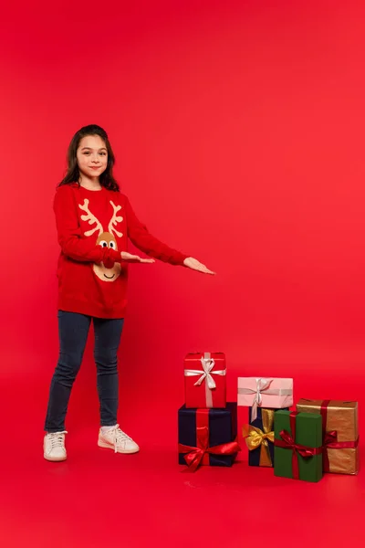 Повна довжина щасливої дитини в в'язаному светрі, вказуючи на різдвяні подарунки на червоному — стокове фото
