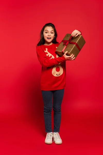 Full length of happy child in knitted sweater holding wrapped Christmas present on red — Stock Photo