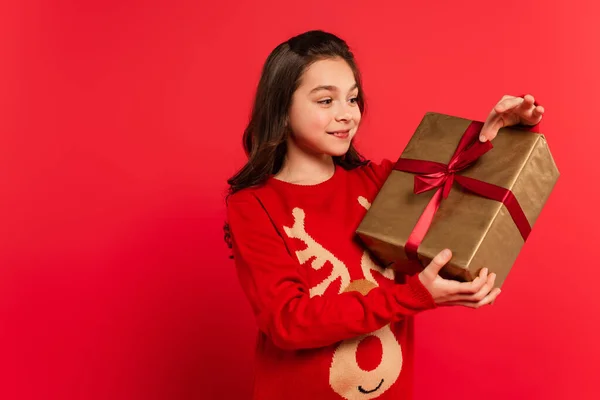 Enfant heureux en pull tricoté regardant enveloppé cadeau de Noël sur rouge — Photo de stock