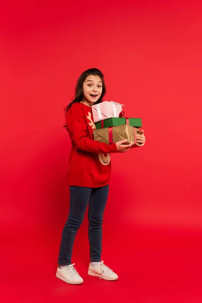Full length of happy girl in winter sweater holding bunch of Christmas presents and smiling on red — Stock Photo