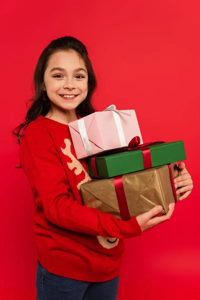 Joyful girl in winter sweater holding bunch of Christmas presents and smiling isolated on red — Stock Photo